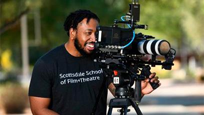 Student looking through a camera and is wearing a black tshirt that reads: Scottsdale School of Film+Theatre in white lettering