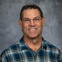 A portrait photo of a white male smiling with brown hair pulled back in a ponytail and wearing glasses. He is wearing a button-down shirt with a collar that has a white, yellow, and blue plaid pattern. The background is varying gray tones.
