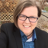 A portrait photo of a white woman smiling with chin-length brown hair, blue framed glasses, and a nose piercing. She is wearing a royal blue button-down shirt with a black blazer. The background includes a light brown lattice fence with fall vines along the top on the left and a rock bed on the right.