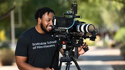 A smiling film school student is looking through camera lens. The student is wearing a Scottsdale Community College t-shirt.