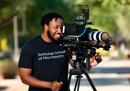 Student looking through a camera and is wearing a black tshirt that reads: Scottsdale School of Film+Theatre in white lettering