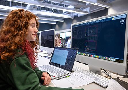 Student with long, curly red hair working on a desktop computer. A laptop is on the desk, along with a paper notepad.