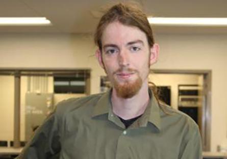 Eli Hallman poses casually in a computer lab; he's dressed in business casual attire.