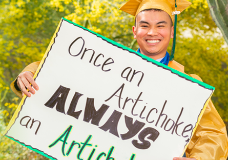 graduate wearing cap and gown holding a sign that reads, Once an Artichoke, Always an Artichoke!