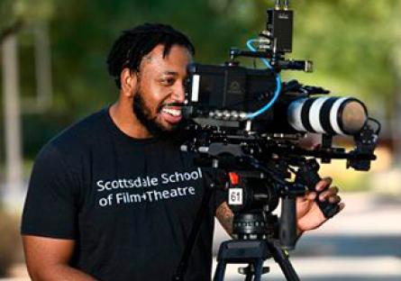 Student looking through a camera and is wearing a black tshirt that reads: Scottsdale School of Film+Theatre in white lettering