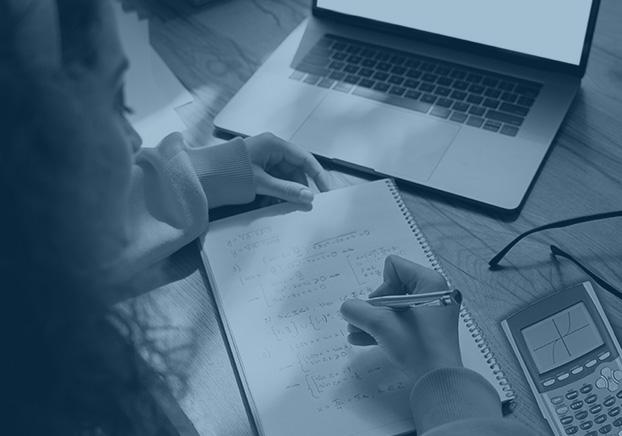 student writing on paper with laptop and calculator on desk