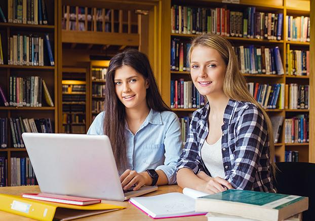 two female college students sitting in a library working together on a laptop